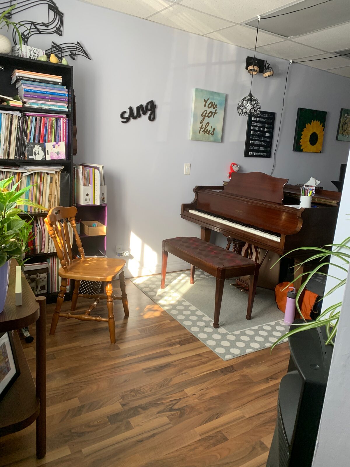 The Alexandria studio: a wooden piano and bench, a bookcase of music, and a “You got this” print on a lilac wall.