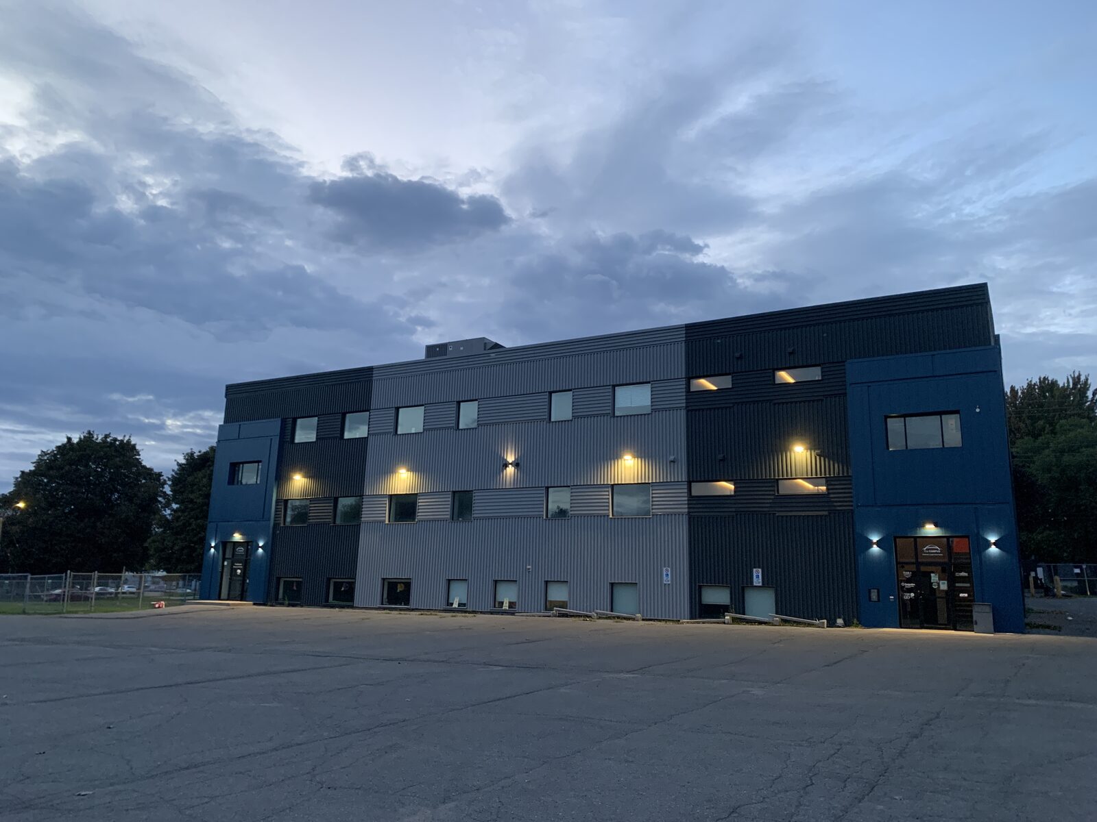 The Cornwall studio building at dusk: three storeys of grey and blue cladding, lit windows, and a parking lot in front.