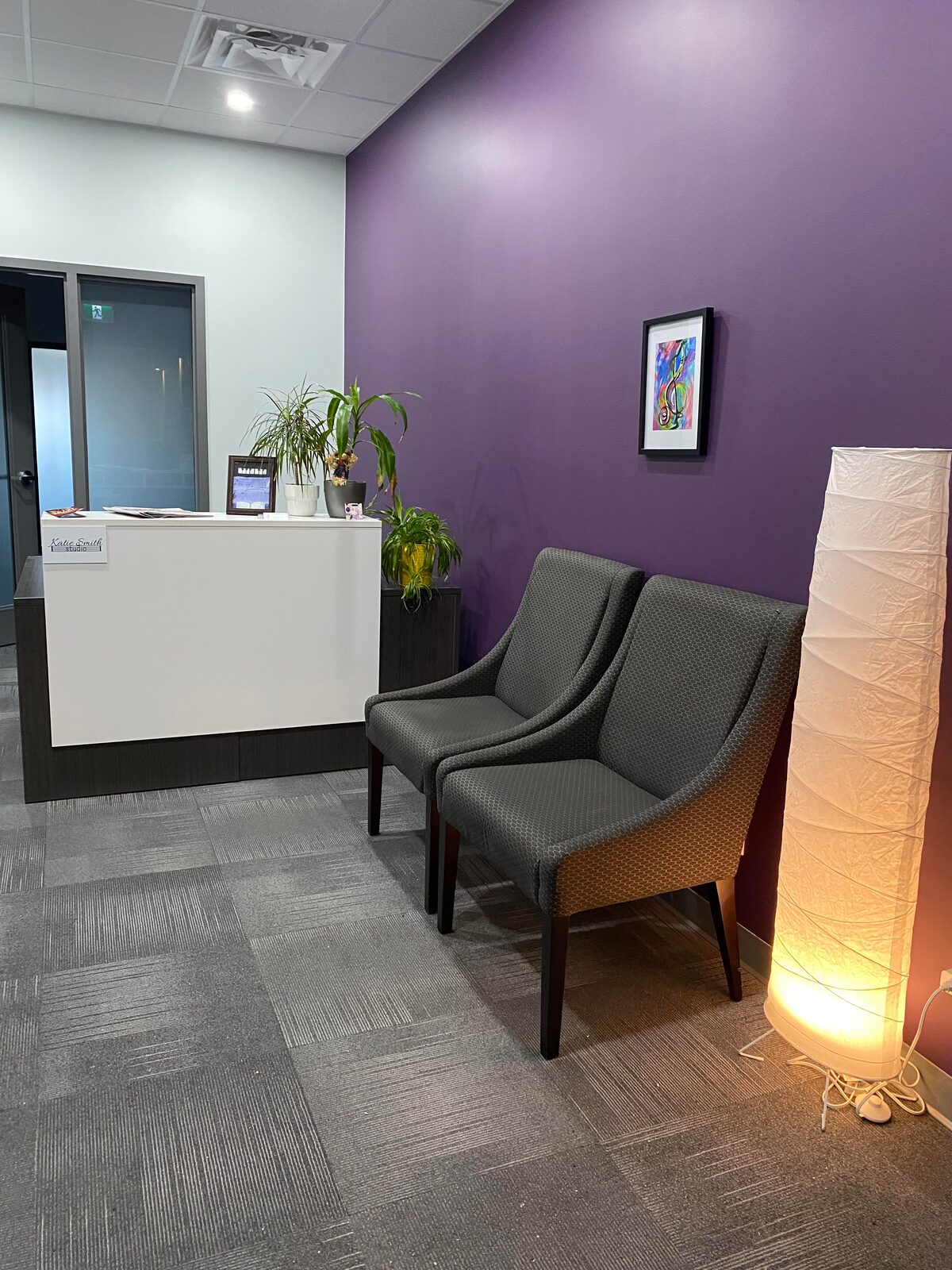 The waiting area of the Cornwall studio: a deep purple wall with a framed treble-clef painting, two grey armchairs, a paper floor lamp, and a reception desk with plants and a Katie Smith studio sign.